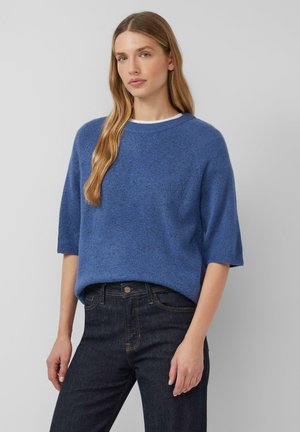 Pull en tricot bleu avec une coupe décontractée, des manches courtes et un col rond, associé à un jean en denim sombre. Texture subtile sur l'ensemble du tissu.