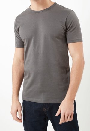Mężczyzna w prostym szarym t-shircie z krótkim rękawem i ciemnoniebieskich dżinsach, stojący na białym tle.
