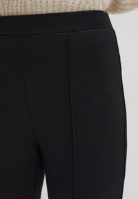 Pantalons noirs extensibles avec une texture lisse, taille haute et détails de couture subtils, présentés sur un fond clair.