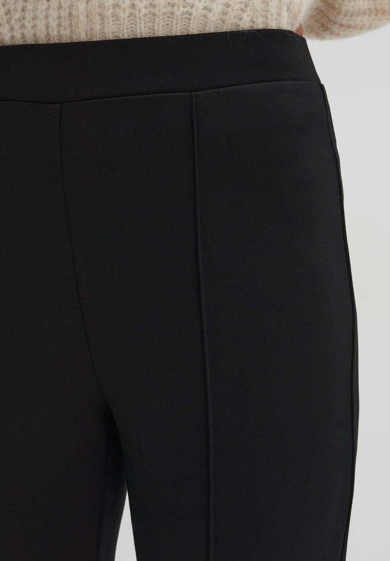 Schwarze dehnbare Hosen mit glatter Textur, hohem Bund und dezenten Nahtdetails, vor einem hellen Hintergrund präsentiert.