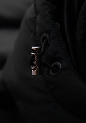 Zwarte pufferjack stof met een close-up van een metalen ritsstop en cordlock, met een gestructureerde oppervlakte en subtiele glans.