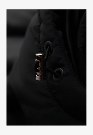 Zwarte pufferjack stof met een close-up van een metalen ritsstop en cordlock, met een gestructureerde oppervlakte en subtiele glans.