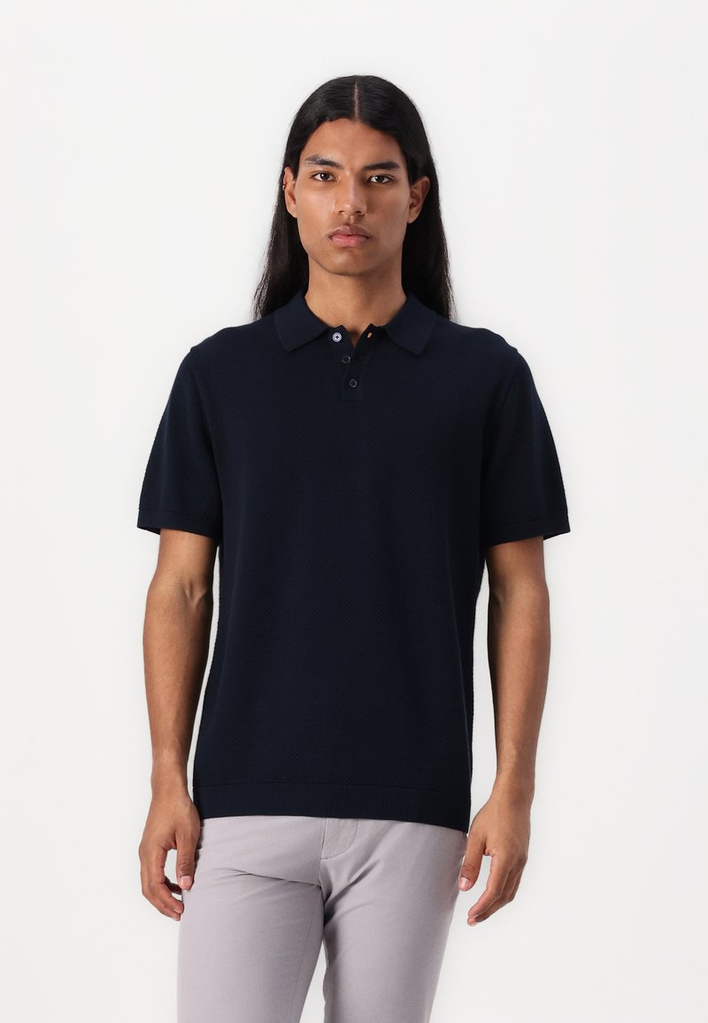 Marineblå kortermet polo-shirt med et teksturert strikkedesign, tre-knapp placket og ribbestrikket krage, kombinert med lysegrå bukser.