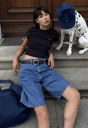 Zwarte geribbelde t-shirt, losse blauwe denim shorts met rafelige uiteinden, zwarte riem met ronde gesp, marineblauwe tas, Dalmatian hond met een blauwe pet.