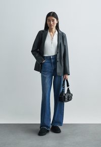 Czarna dopasowana marynarka, biały prążkowany top ze sznurowanym detalem, dzwony w kolorze niebieskim, czarna mini torba i czarne zamszowe buty. Neutralne tło.