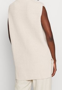 Béžový oversized svetřík bez rukávů s žebrovanou strukturou, bočními rozparkami a vysokým límcem. Měkký pletený materiál s volným střihem.