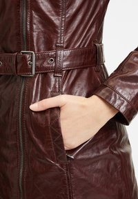 Cappotto in pelle bordeaux con vita cintrata, chiusura con zip e tasche laterali; superficie texture con dettagli lucidi discreti.