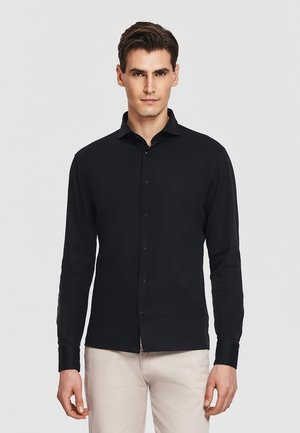 Mężczyzna w dopasowanej czarnej koszuli na guziki z długim rękawem i jasnobeżowych spodniach, stojący na tle jednolitego jasnego tła.