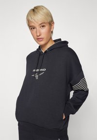 Marineblå hettegenser laget av bomullsblanding, med en frontlomme, hvit logo ogstripete detaljer på ermene.