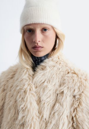 Beige faux fur coat with a long, shaggy texture. Worn over a dark striped turtleneck. Model also wearing a white ribbed knit beanie.