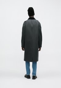 Lang mørkegrå frakke med notch-krave, med en lige snit. Båret over lyseblå jeans og sorte sko, set bagfra.