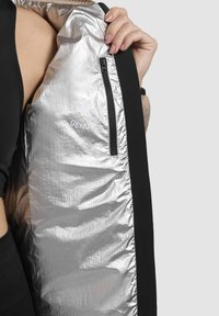 Giacca a vento nera e argento con tasca con zip; tessuto metallico lucido con texture arricciata; logo stampato in bianco.