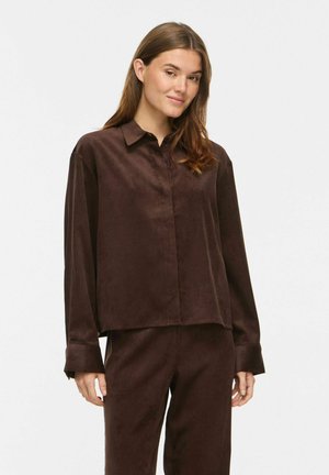 Jeune femme aux longs cheveux bruns portant une chemise amples marron foncé boutonnée et un pantalon assorti sur un fond clair uni.