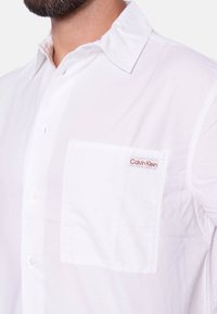 Camisa blanca de manga corta con cuello abotonado, que presenta un bolsillo en el pecho izquierdo y un pequeño logo de Calvin Klein en rojo. Tejido suave y liviano.