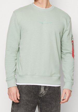 Hombre con sudadera verde claro con pequeño logo y bolsillo con cremallera y etiqueta roja en la manga izquierda, combinado con vaqueros gris oscuro.