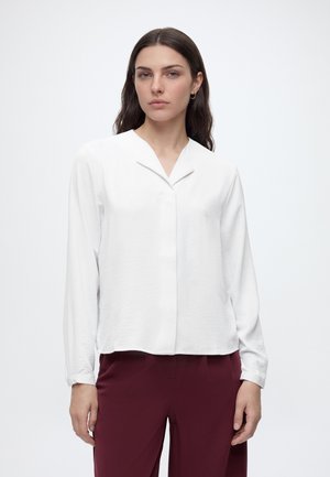 Frau mit langen dunklen Haaren, die eine weiße Bluse mit langen Ärmeln und bordeauxrote Hose trägt, steht vor einem einfarbigen hellen Hintergrund.