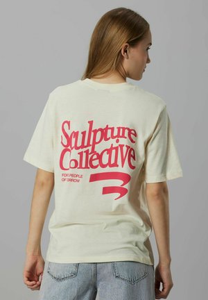 Kvinde med langt hår iført en cremefarvet skjorte med rød tekst "Sculpture Collective" sammen med lyseblå jeans, stående foran en grå baggrund.