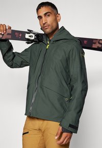 Chaqueta impermeable de color verde oscuro con capucha, cierre de cremallera y puños ajustables. Presenta detalles amarillos y múltiples bolsillos.