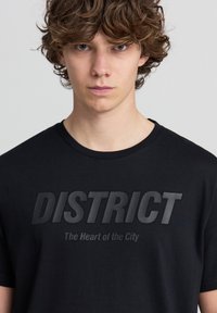 T-shirt in cotone nero con grande e audace scritta "DISTRICT" in finitura opaca, accompagnata dalla frase secondaria "Il Cuore della Città" sottostante.