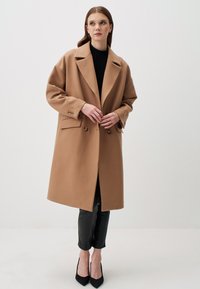 Cappotto di cammello con colletto a punta, frontale doppiopetto e due bottoni. Silhouette strutturata, abbinata a pantaloni neri e tacchi.