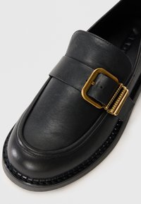 Black leather loafers featuring a wide strap and a gold rectangular buckle. The shoe has a rounded toe and a textured sole.