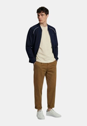 Uomo con giacca blu navy con zip, t-shirt beige, pantaloni marroni e scarpe da ginnastica bianche, in piedi con le mani in tasca, guarda verso sinistra.