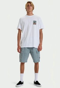 Young man standing, wearing a white graphic t-shirt, light blue cargo shorts, white socks, and black slip-on shoes.