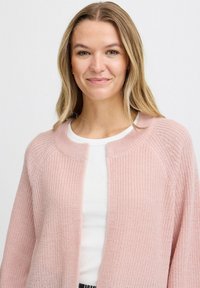 Šviesiai rožinė atvira priekyje džemperis, tekstūruota mezginys ir apvalus kaklas. Dėvimas virš balto viršaus, su įstrižais akcentais aplink kaklą.