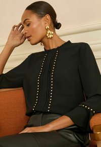 Moteris su atgal sušukuotais plaukais, dėvinti didelius auksinius auskarus ir juodą palaidinę su auksiniais širdies formos smeigtukais, sėdinti ant oranžinės kėdės.