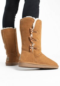 Stivali alti in suede color tan con morbido rivestimento bianco, quattro dettagli triangolari con cordini in legno e suole in gomma testurizzata per una maggiore aderenza.