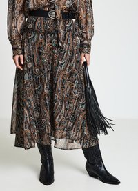 Jupe longue noire et marron à motif paisley, avec une taille cintrée et une silhouette fluide, associée à un sac à franges en cuir noir et des bottes.