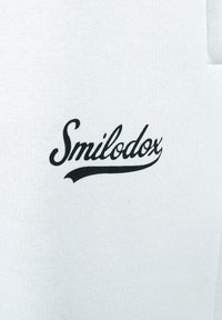 Tejido de algodón blanco con un destacado logo negro de "Smilodox" en una fuente cursiva, presentando una textura suave y un diseño limpio.
