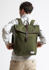 Mochila de lona verde con diseño de rollo, correa y cremallera de cuero marrón, bolsillo frontal y correas de hombro ajustables.