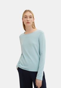 Light blue long-sleeve shirt in a textured knit fabric, featuring a round neckline and a relaxed fit. Paired with dark pants.