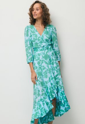 Kobieta o kręconych włosach, ubrana w niebiesko-zieloną sukienkę kopertową w kwiaty z falbaną u dołu, stojąca na jednolitym białym tle.