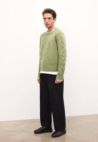 Hele rohelise värvi kudumist-sweater koos kaablipatternitega, ribaga kaeluse ja äärega, koos mustade laia jalaga pükste ja mustade tennisetossudega.