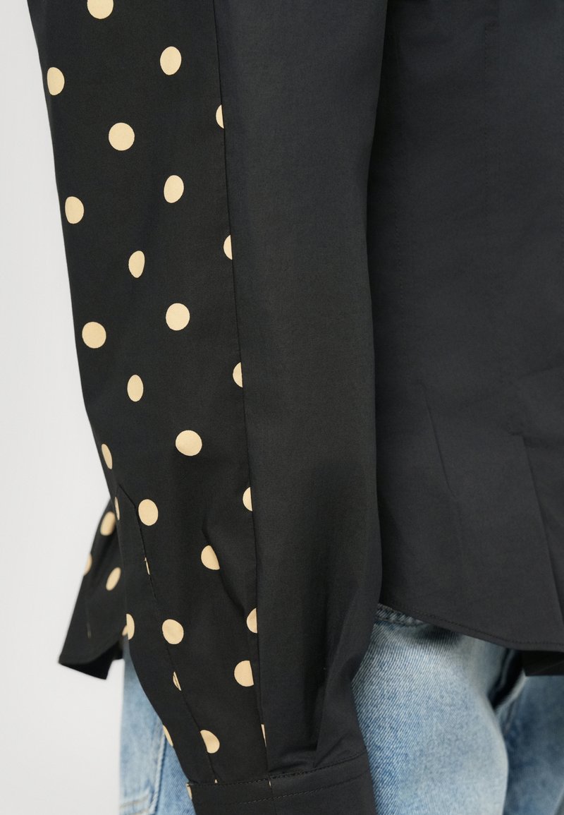Zwarte longsleeve met beige stippen op één mouw, volledig zwart aan de andere kant, gedragen over lichtblauwe denim jeans.