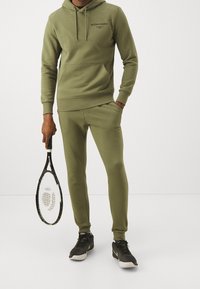 Sudadera verde oliva y pantalones de chándal a juego, hechos de tejido suave. La sudadera presenta un bolsillo canguro y un logo; los pantalones tienen un corte ajustado. Sosteniendo una raqueta de tenis.