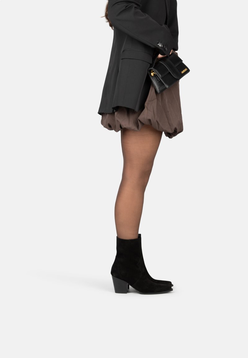 Botines de ante negro con punta afilada, tacón alto y grueso, y textura suave, combinados con una falda corta marrón y una chaqueta negra.