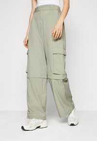 Αρκετά χαλαρές ανοιχτοπράσινες cargo παντελόνες με μεγάλες πλαϊνές τσέπες και λευκά sneakers, φορεμένες από ένα άτομο που στέκεται σε ένα απλό φόντο.