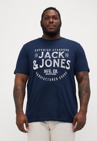 Námořnická modrá bavlněná tričko s velkým bílým textem s názvem značky "JACK & JONES" a další typografií. Krátké rukávy, kulatý výstřih.