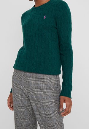 Person wearing a dark green cable-knit sweater and gray plaid pants, standing against a plain light gray background.