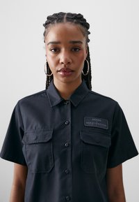 Black short-sleeve shirt with button front, two chest pockets, and a branded patch reading "Dickies x Harley-Davidson." Smooth texture.
