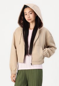 Veste croppée beige avec capuche, poignets élastiques et poche avant. Portée par-dessus un pull rose pâle et un pantalon plissé olive.