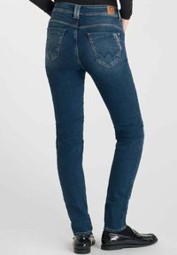 Jeans skinny en denim bleu foncé, avec une taille mi-haute, deux poches arrière avec un motif de couture subtil, et une texture lisse.