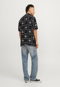 Camisa negra de manga corta con un patrón de hojas de palma blancas, combinada con unos vaqueros anchos de color azul claro y zapatillas negras. Vista lateral, postura de pie.