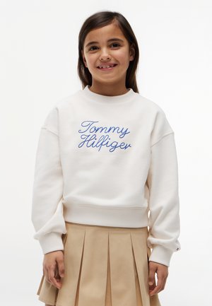 Smilende pige med langt brunt hår iført hvid Tommy Hilfiger sweatshirt og beige plisseret nederdel mod ensfarvet baggrund.
