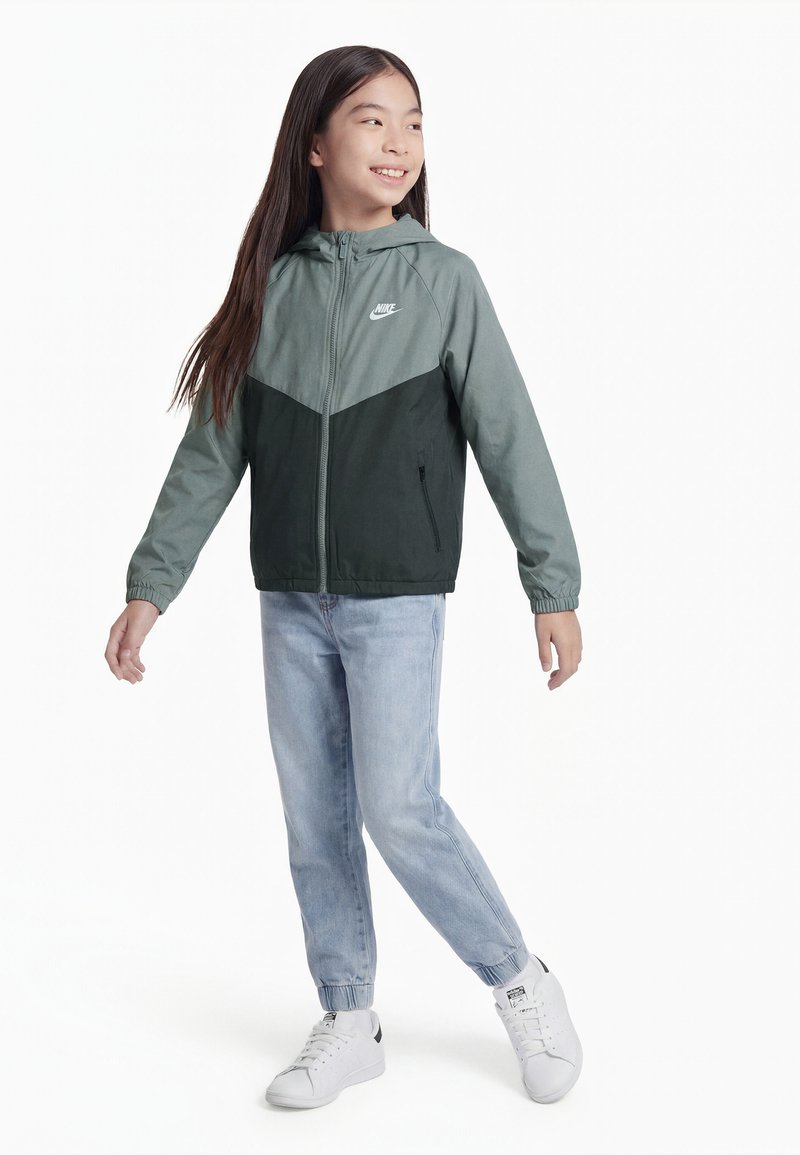 Fille souriante, debout avec les bras légèrement écartés, portant une veste Nike bicolore verte, un jean bleu clair et des baskets blanches sur fond blanc.