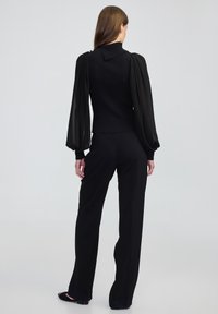 Top nero a coste con maniche a sbuffo trasparenti, caratterizzato da un colletto alto e dettagli con bottoni. Abbinato a pantaloni neri a vita alta e ballerine.