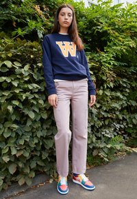 Joven mujer vistiendo sudadera azul marino con una gran "W", pantalones rosa claro y zapatillas coloridas, se encuentra al aire libre frente a un denso follaje verde.